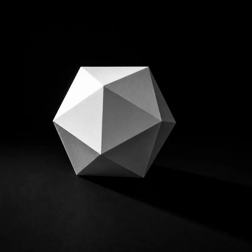 Icosaèdre en papier blanc plié posé sur une surface noire, éclairé en lumière rasante depuis le haut à gauche