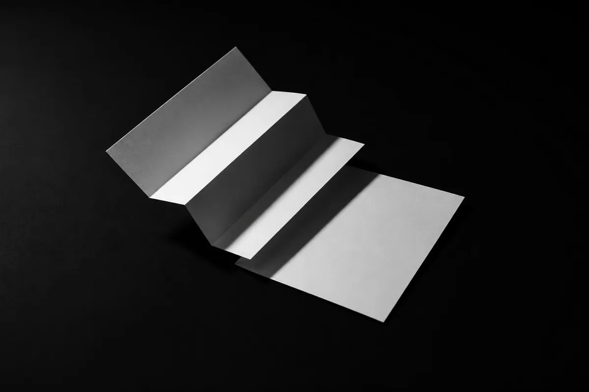 Feuille de papier blanc à moitié dépliée : une moitié garde des plis nets, l’autre est lisse, en lumière rasante sur fond noir